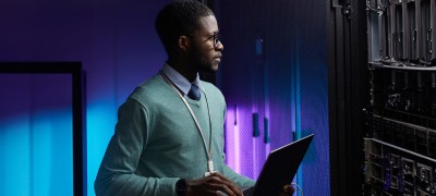 A man stands in a server room, holding a laptop.