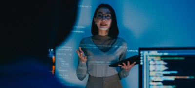 A woman stands with a tablet and a computer code is displayed on a monitor behind her.